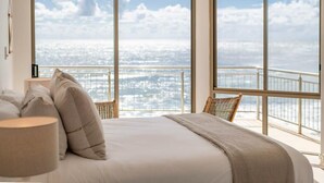 A bedroom with a large bed, two chairs, and a view of the city through large windows. - Oceans 8 - Oceanstays -Absolute Oceanfront with Fireplace (Yamba)