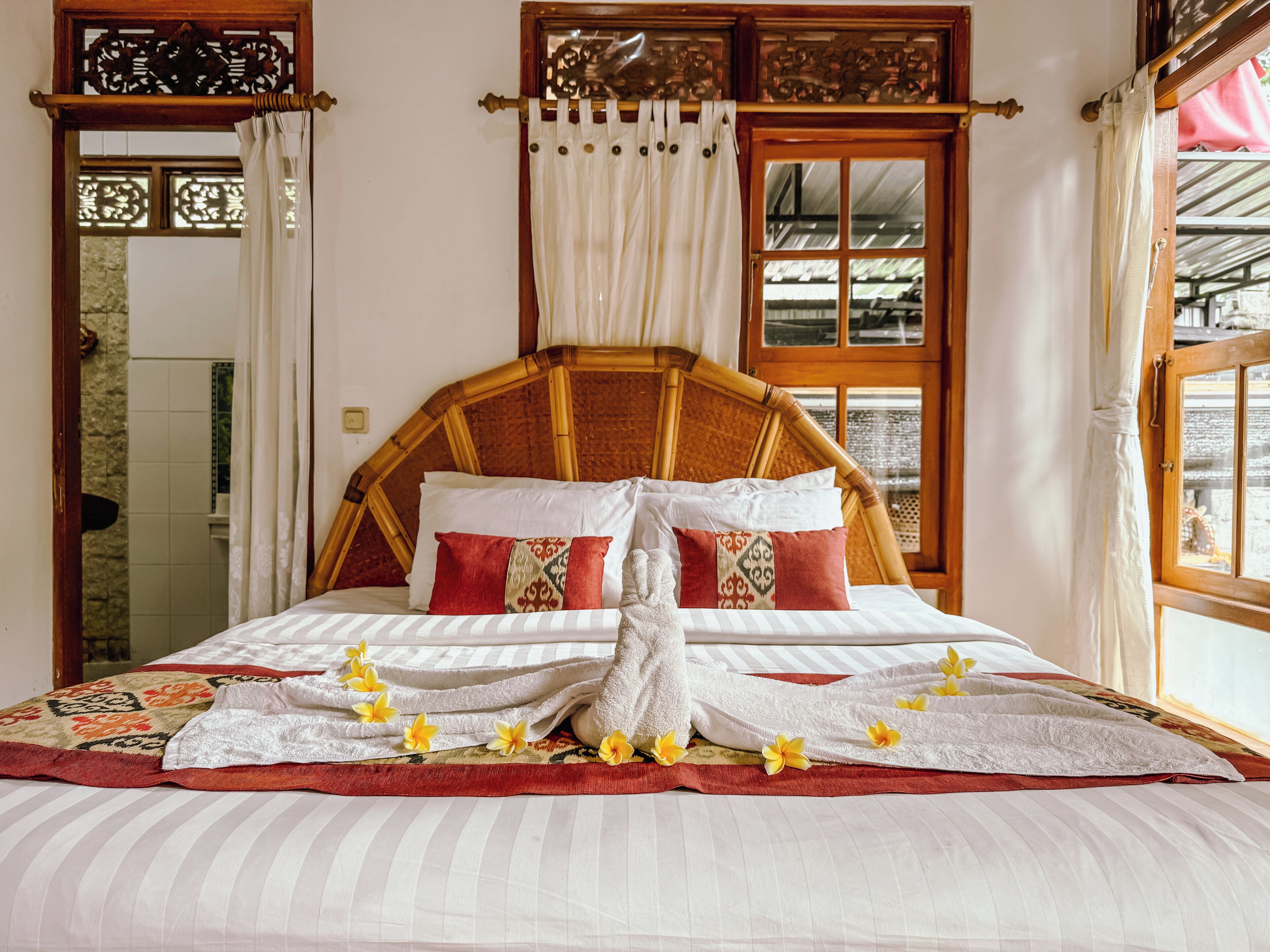 Una cama bien hecha con ropa de cama blanca y a rayas, almohadas rojas y estampadas, y una toalla con flores amarillas.