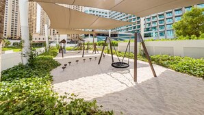 A playground with swings and a slide, surrounded by greenery and buildings. - Luxurious 1BR Oasis Panoramic Views (Dubai)