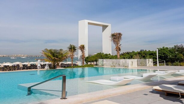 A pool area with a modern white structure, lounge chairs, and palm trees. - Luxurious 1BR Oasis Panoramic Views (Dubai)