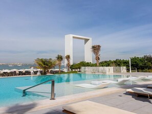 A pool area with a modern white structure, lounge chairs, and palm trees. - Luxurious 1BR Oasis Panoramic Views (Dubai)