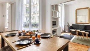 Un comedor con una mesa de madera preparada para el desayuno, una ventana con cortinas, un sofá negro y un espejo en la pared.