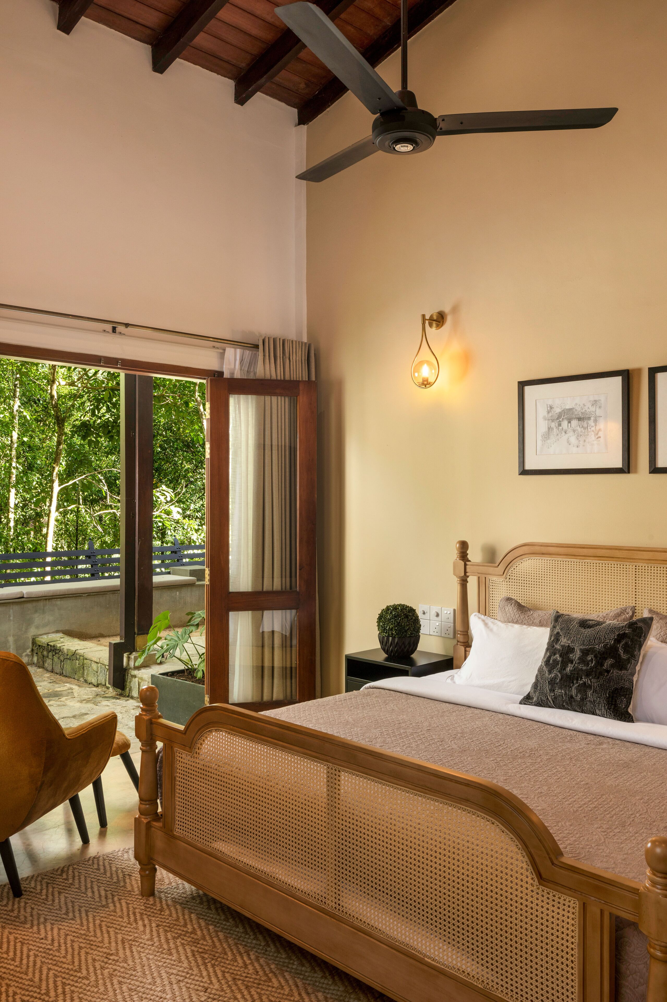 A bedroom with a bed, a chair, a ceiling fan, a window with a view of trees, and a wall-mounted light fixture.