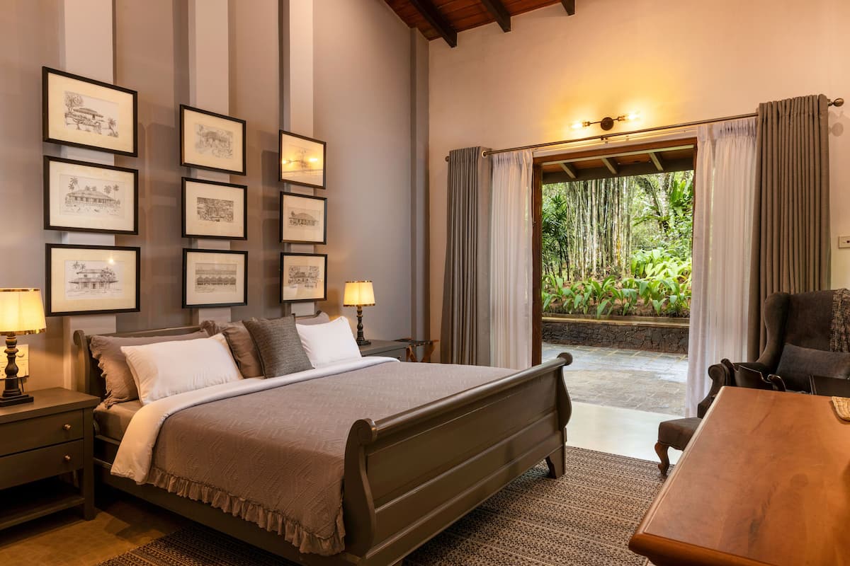 A bedroom with a large bed, bedside tables with lamps, framed artwork on the wall, and a view of greenery through a door.