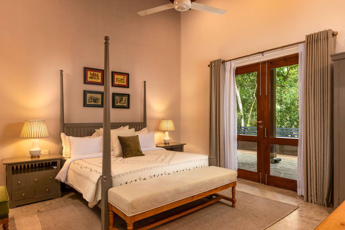 A four-poster bed with a bench, bedside tables with lamps, and a view of a garden through a glass door.