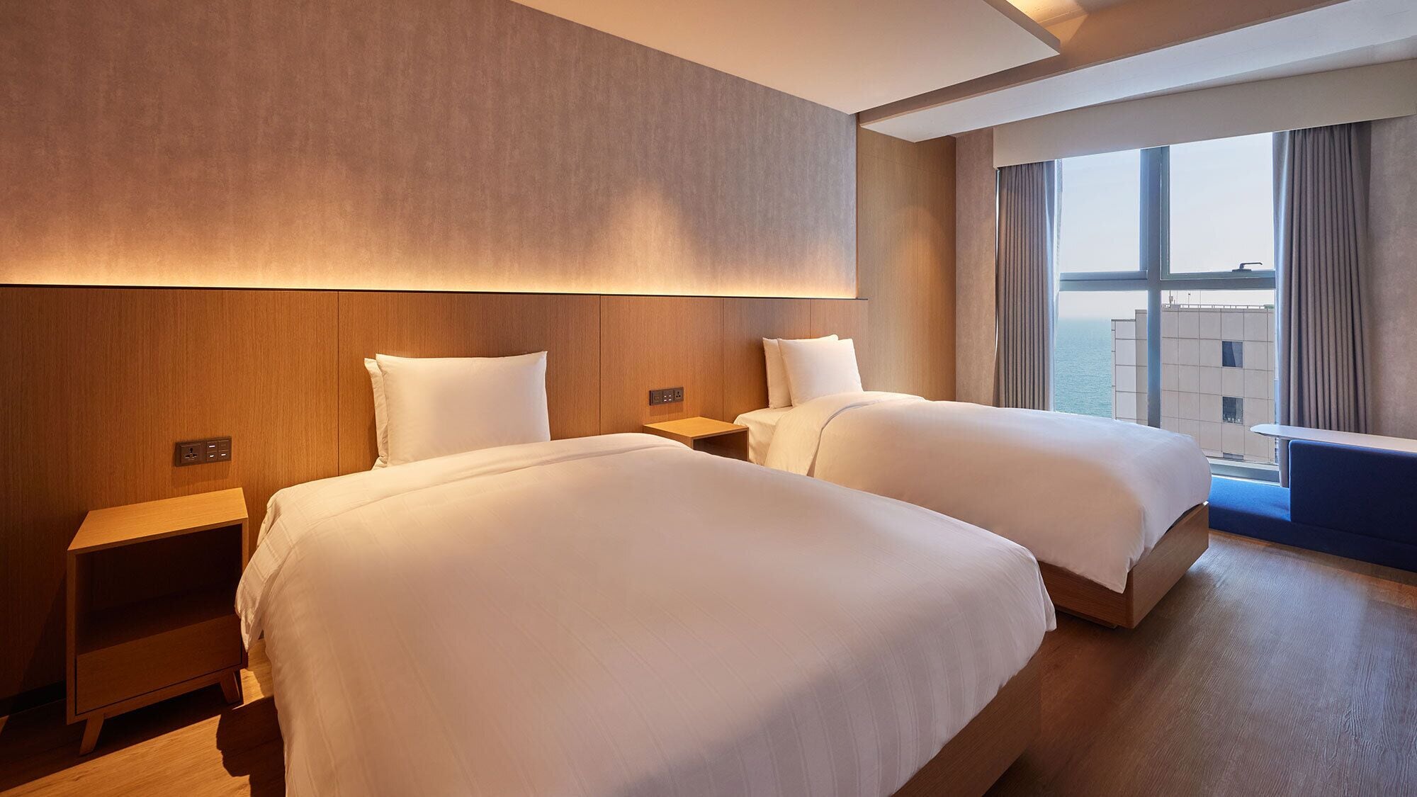 A hotel room with two beds, wooden headboards, a nightstand, and a large window offering a view of the city.