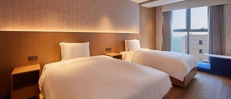 A hotel room with two beds, wooden headboards, a nightstand, and a large window offering a view of the city.
