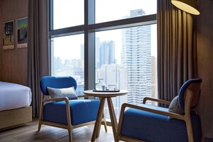 Habitación de hotel moderna con un ventanal grande, una cama, dos sillones azules y una pequeña mesa redonda con una jarra de vidrio y un vaso de agua.