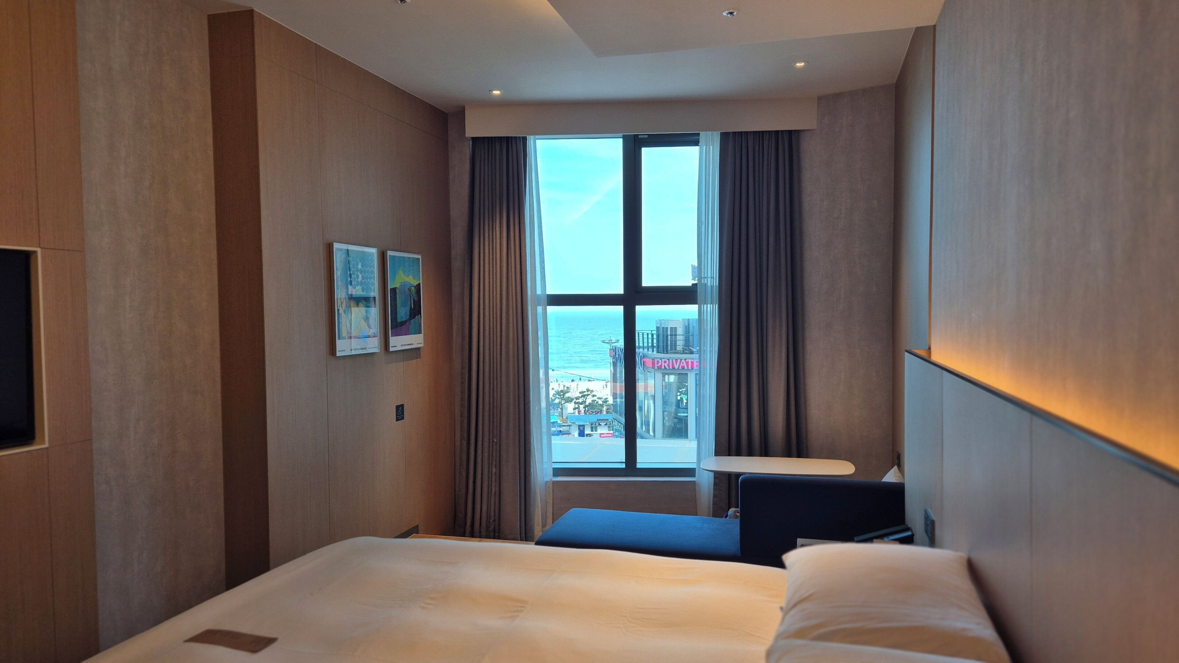A hotel room with a large bed, a small desk, and a view of the city through a large window.