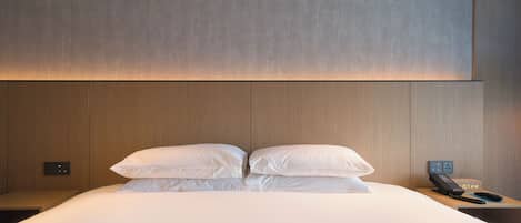 A neatly made bed with white linens and pillows, a bedside table with a phone and a clock, and a wall-mounted light fixture.