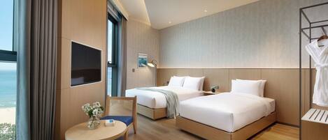 A modern hotel room with two beds, a TV, a desk, and a view of the beach.