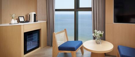 Una camera d'albergo moderna con pavimento in legno, una zona pranzo con sedute imbottite blu, un tavolino, una macchina da caffè, una TV a muro e vista sul mare attraverso ampie finestre.