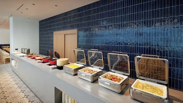 Disposição de buffet com diversos recipientes de comida em um balcão, piso estampado e parede com azulejos azuis.