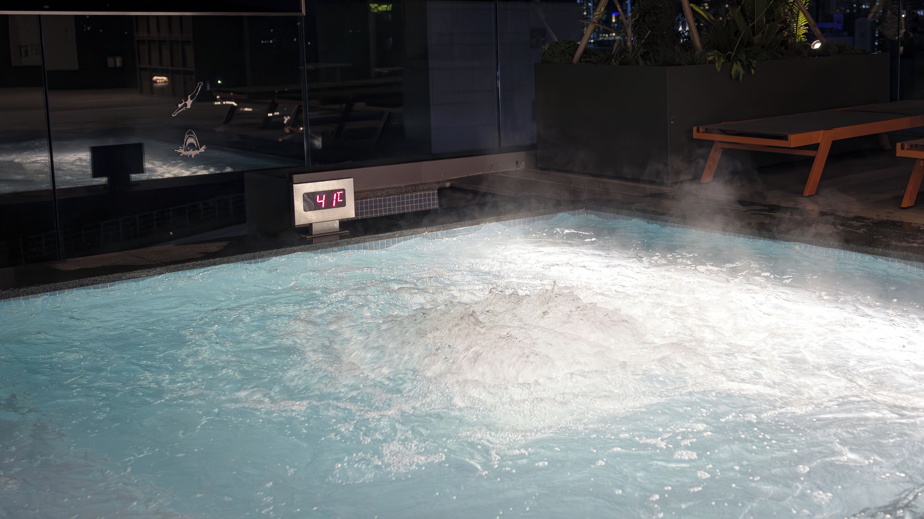 A hot tub with a digital temperature display, surrounded by a glass enclosure and a wooden bench.