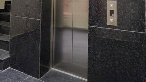 Un ascensor con acabado metálico, ubicado en un edificio moderno con paredes de mármol oscuro y piso de baldosas.