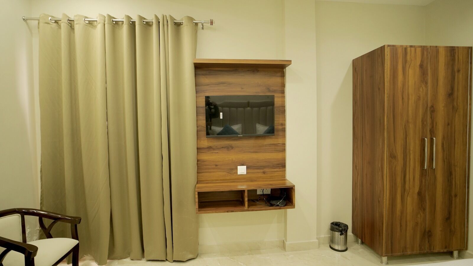 A room with a wooden cabinet, a television mounted on a wooden shelf, a chair, and a curtain.