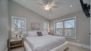 Un dormitorio con cama, dos mesitas de noche, ventilador de techo y ventanas amplias.