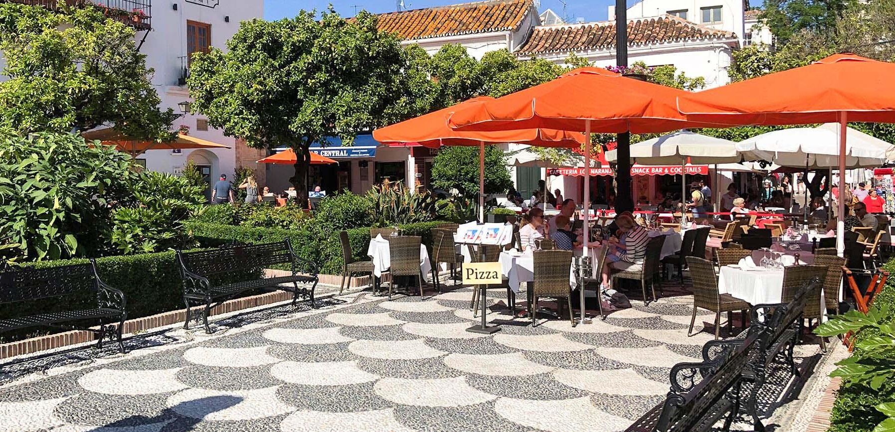 An outdoor café with tables and chairs under orange umbrellas, surrounded by greenery and buildings.