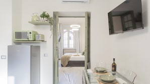 Una cuina moderna amb nevera, microones i torradora a les estantes, una zona de menjador amb taula de vidre i cadires, i un televisor fixat a la paret.