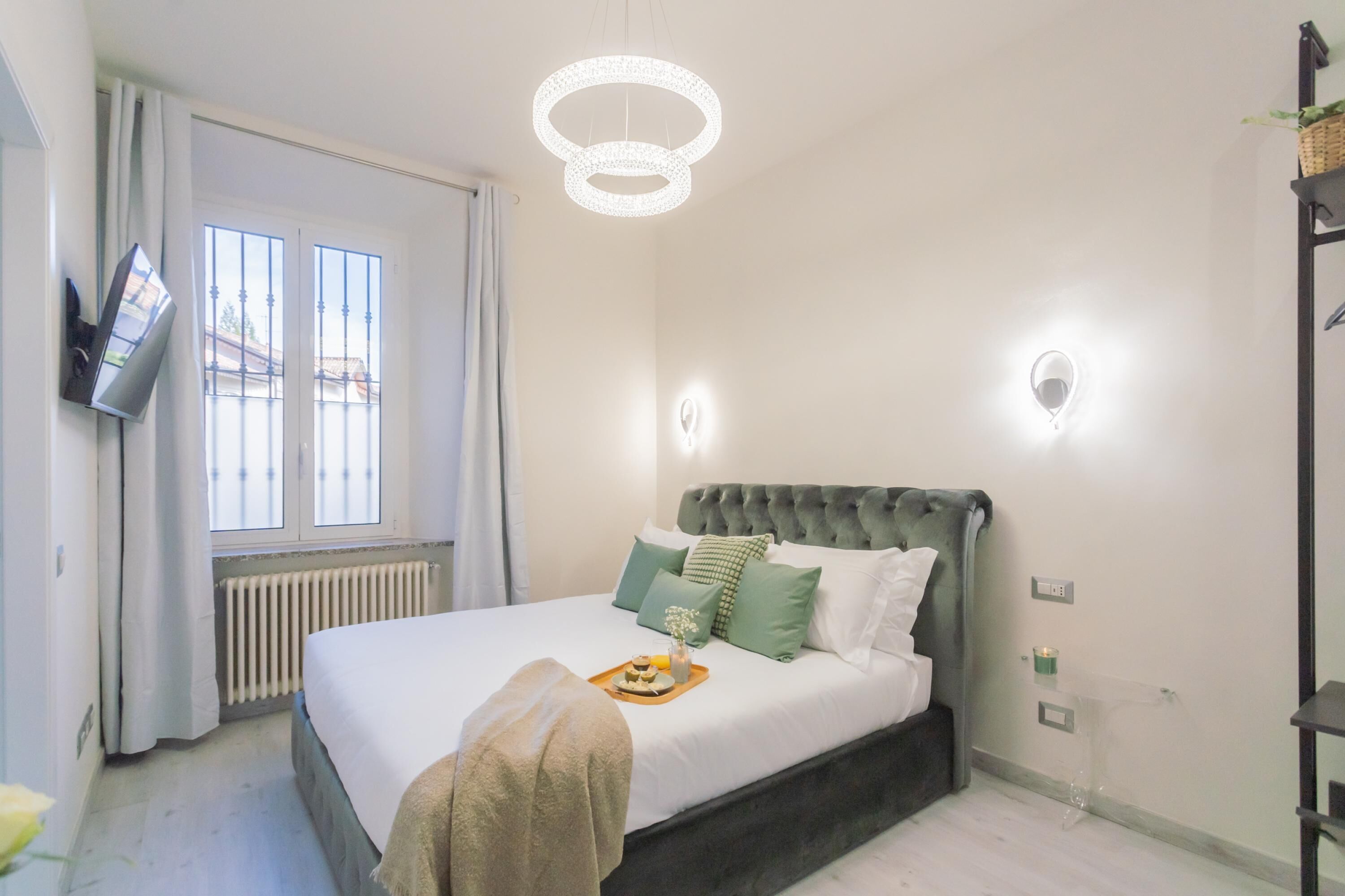 A bedroom with a bed, a TV mounted on the wall, a window with curtains, a radiator, and a chandelier.