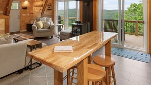 Interior de una cabaña de madera con comedor, sofá y un amplio ventanal con vistas a un bosque.