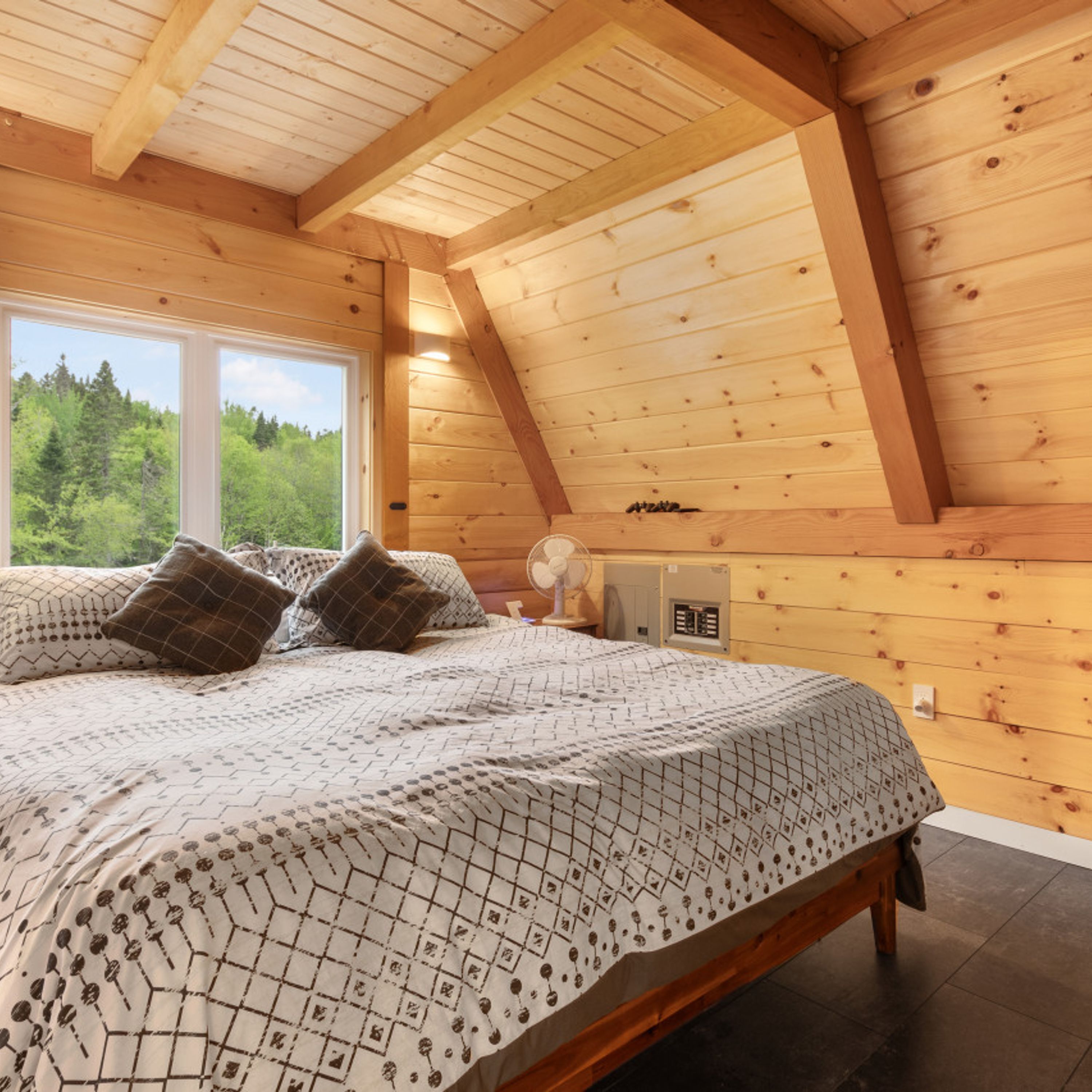 Une chambre à coucher en bois, avec un lit, deux oreillers, une table de chevet et une fenêtre donnant sur des arbres.