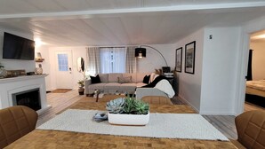 A living room with a sofa, coffee table, and a potted plant. There is a fireplace, a television, and a bed in the background.