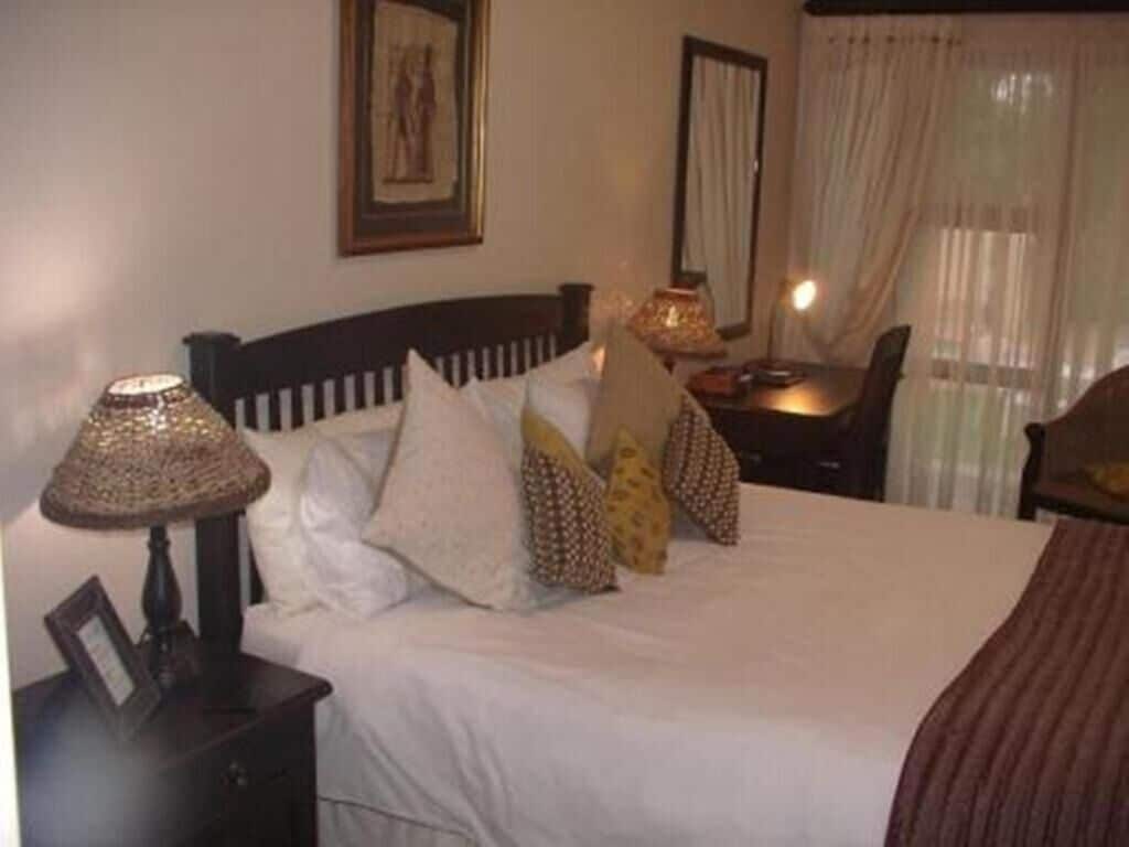 Un lit bien fait, avec du linge de lit blanc et des coussins à motifs, une lampe de chevet, un tableau encadré et une petite table avec une lampe.