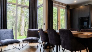 Et moderne spiseområde med et træbord og mørke stole, store vinduer med gardiner, og en indbygget ovn i baggrunden.