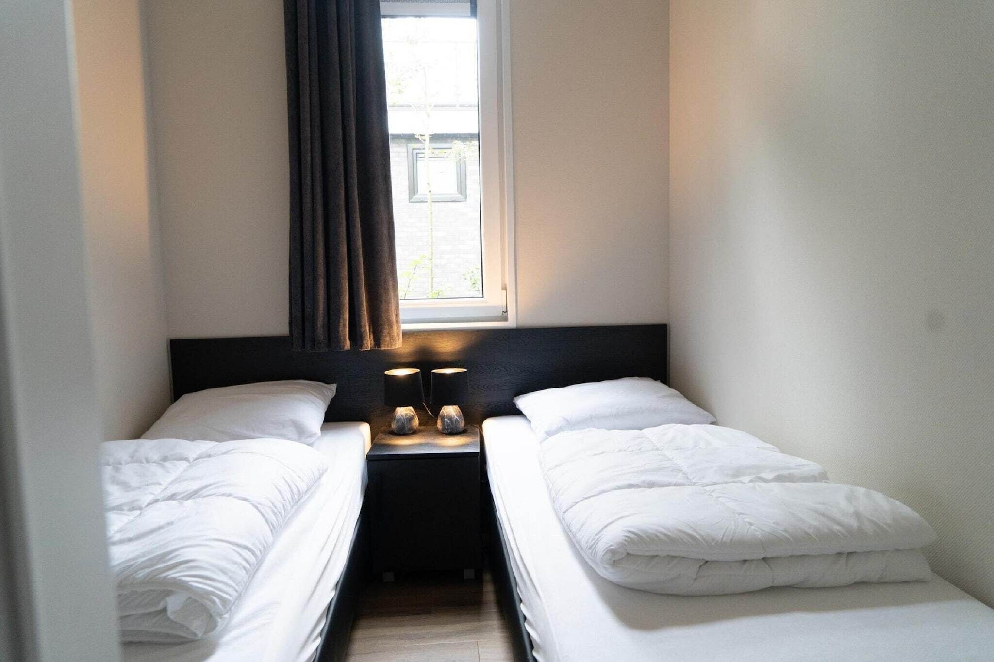 A small, clean room with two single beds, a nightstand with a lamp, and a window with curtains.