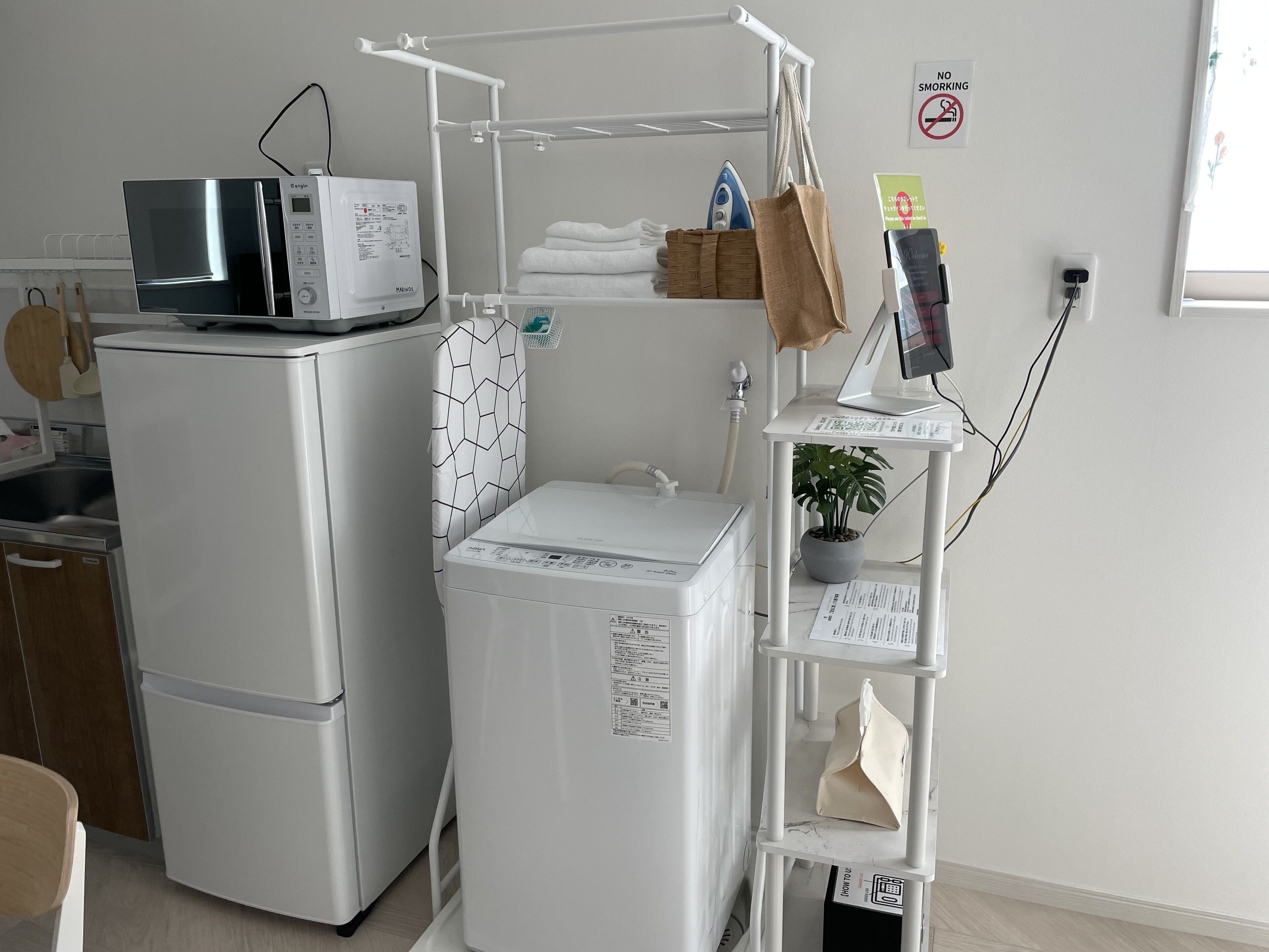 A compact kitchen with a refrigerator, microwave, and washing machine. There is a drying rack, a shelf with a plant, and a 'No Smoking' sign on the wall.