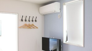Ein Hotelzimmer mit Flachbildfernseher an der Wand, einem Fenster mit weißem Rollo, einer Wandklimaanlage und einem Kleiderständer mit Kleiderbügeln.
