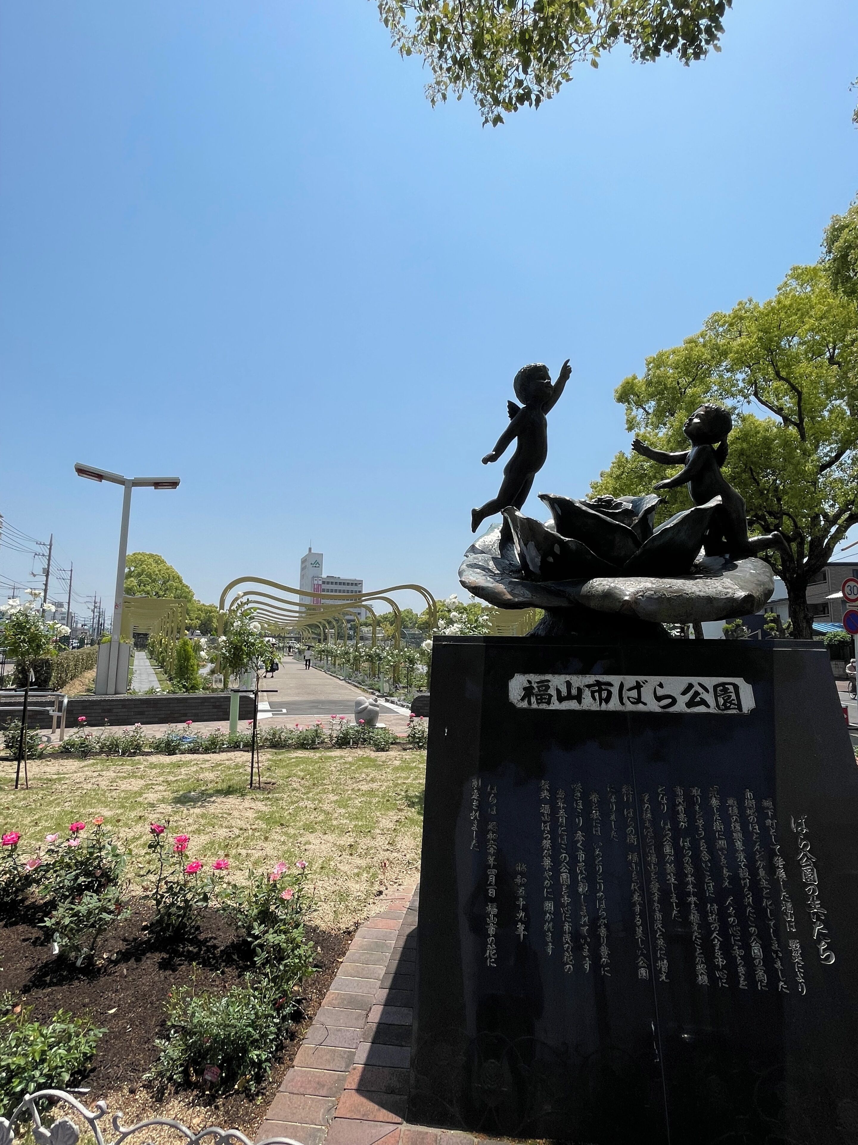 A park with a statue of a child on a rock, surrounded by flowers and a clear sky.