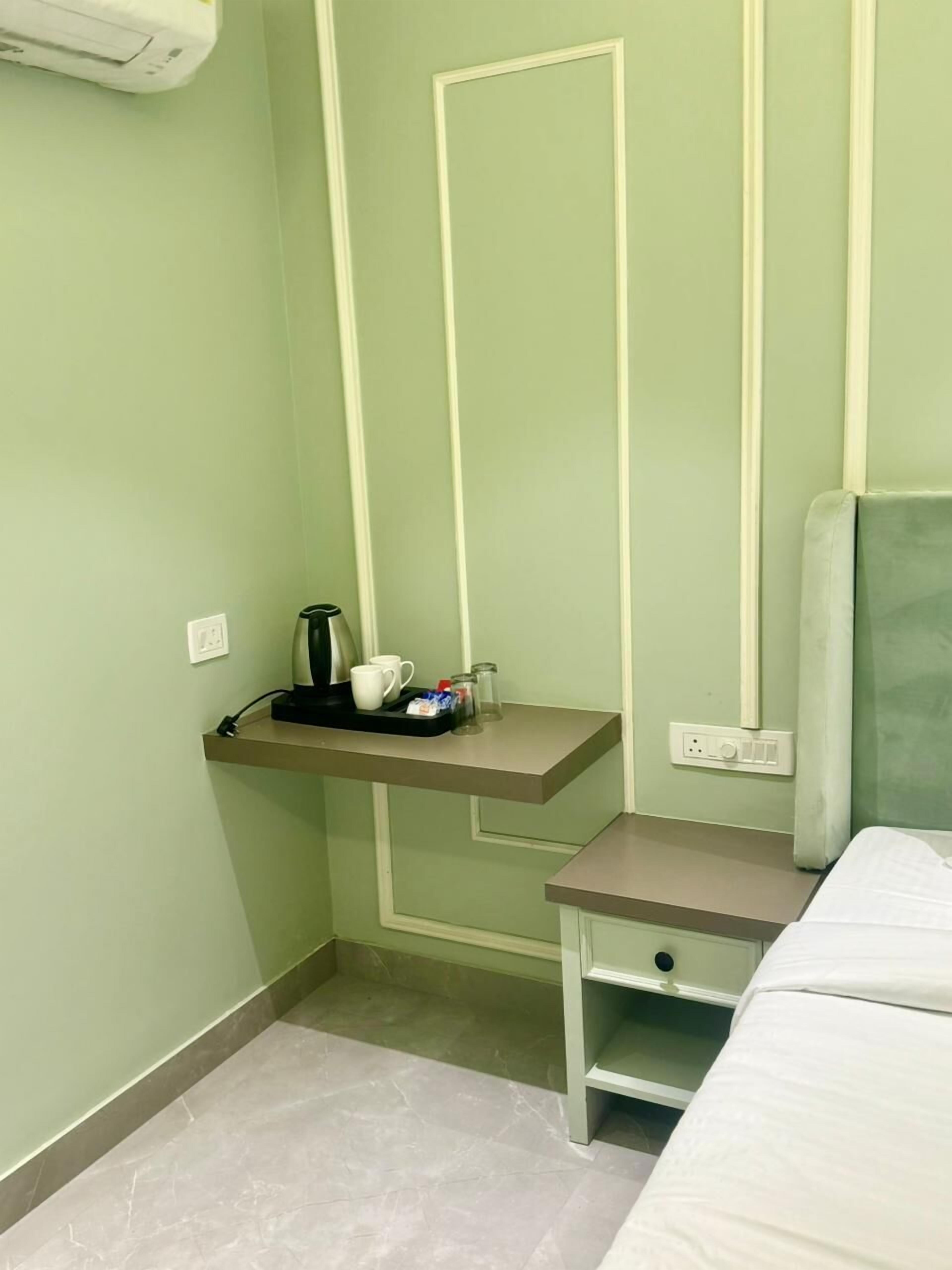 Une petite pièce, bien rangée, comprenant un lit, une table de chevet, une étagère avec une bouilloire, des tasses et des condiments, ainsi qu’un climatiseur fixé au mur.