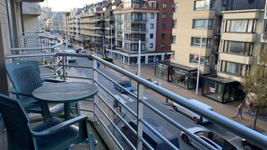 Un balcon avec des sièges pour l’extérieur, une vue sur une rue de la ville et des immeubles abritant des boutiques et des appartements.