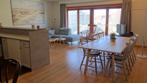 A modern living room with a sofa, dining table, and chairs, a kitchen area, and a large window with a city view. - Apartment in Nieuwpoort near the Beach (Nieuwpoort)