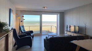 A modern living room with a large sliding glass door leading to a balcony with a view of the beach. There are two black leather chairs, a dark patterned sofa, and a small table with a lamp. A flat-screen TV is mounted on the wall, and there is a plant on the left side. - Apartment in Nieuwpoort by the Marina (Nieuwpoort)