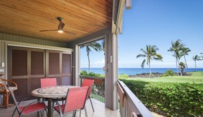 Ein überdachter Außenbereich mit Tisch und Stühlen, einem Deckenventilator und Blick auf das Meer und Palmen.