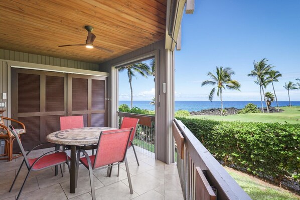 Ein überdachter Außenbereich mit Tisch und Stühlen, einem Deckenventilator und Blick auf das Meer und Palmen.