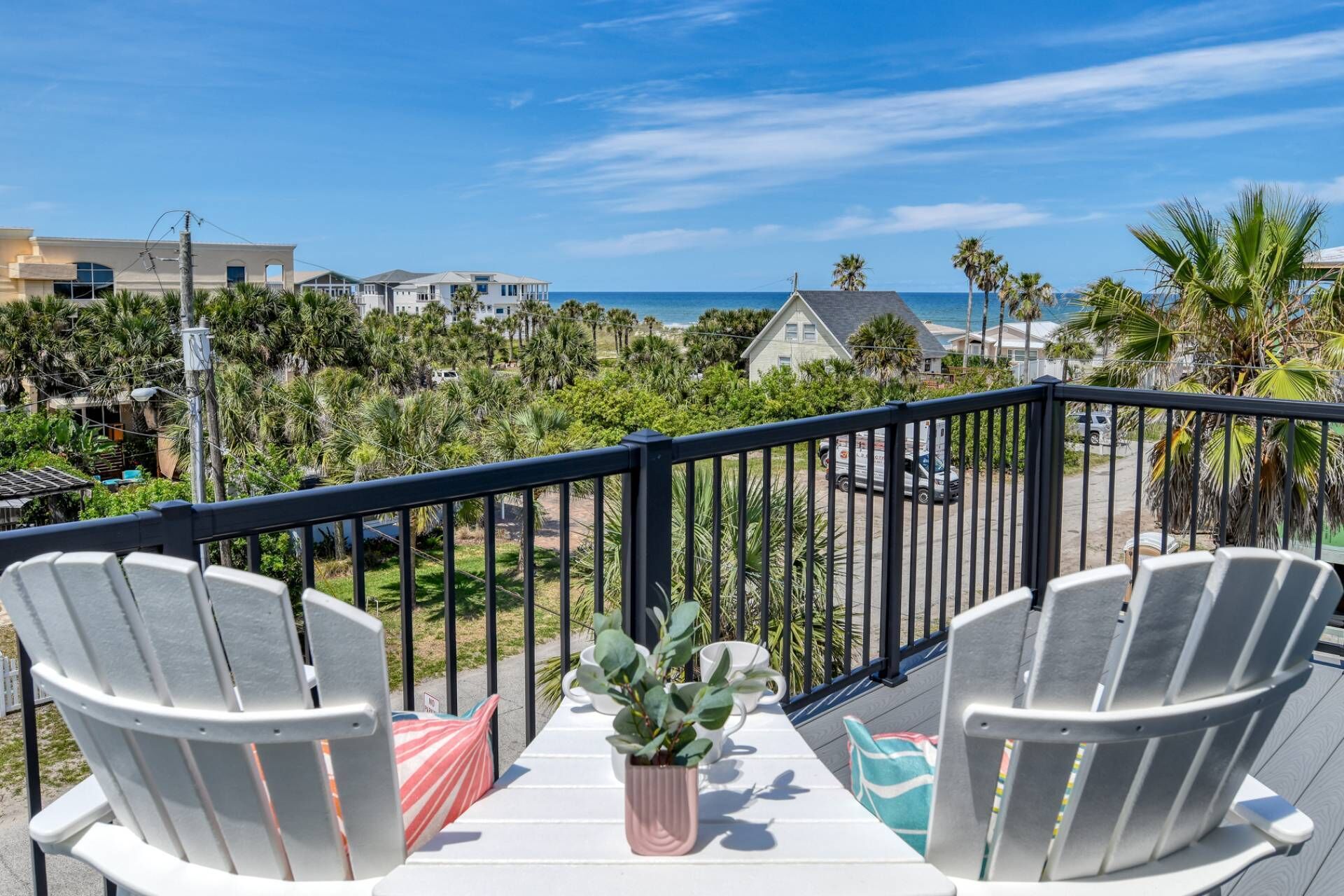 A balcony with two Adirondack chairs, a small table, and a potted plant. The view includes a beach and residential buildings.