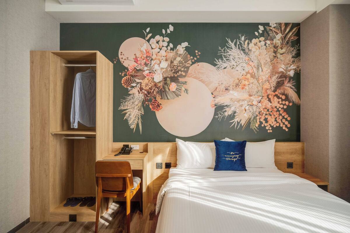 A hotel room with a wooden wardrobe, a desk with a telephone, a bed with white bedding and a blue pillow, and a wall mural with floral and circular designs.
