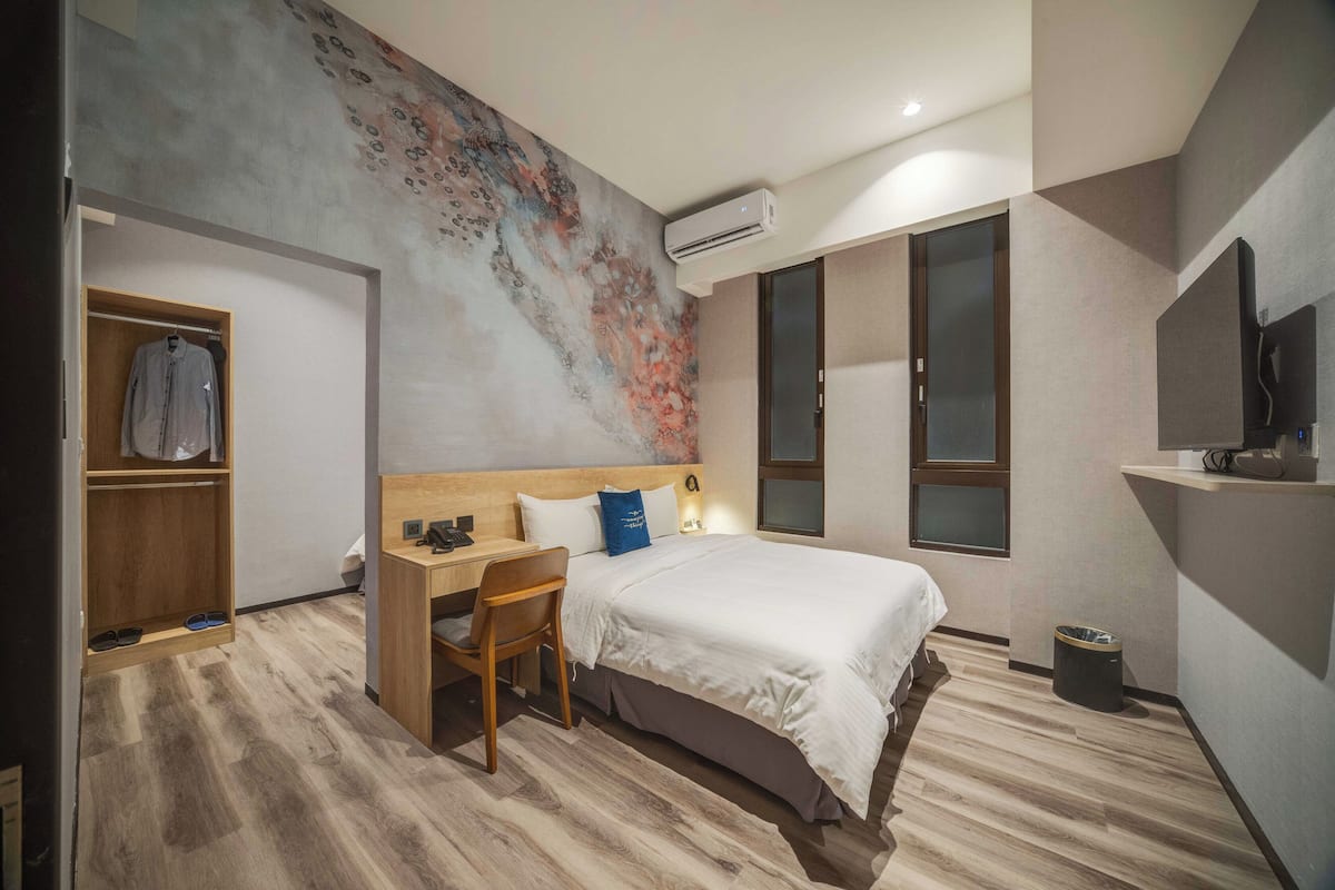 A modern bedroom with a large bed, a wooden desk, a chair, a wardrobe, and a large abstract painting on the wall.