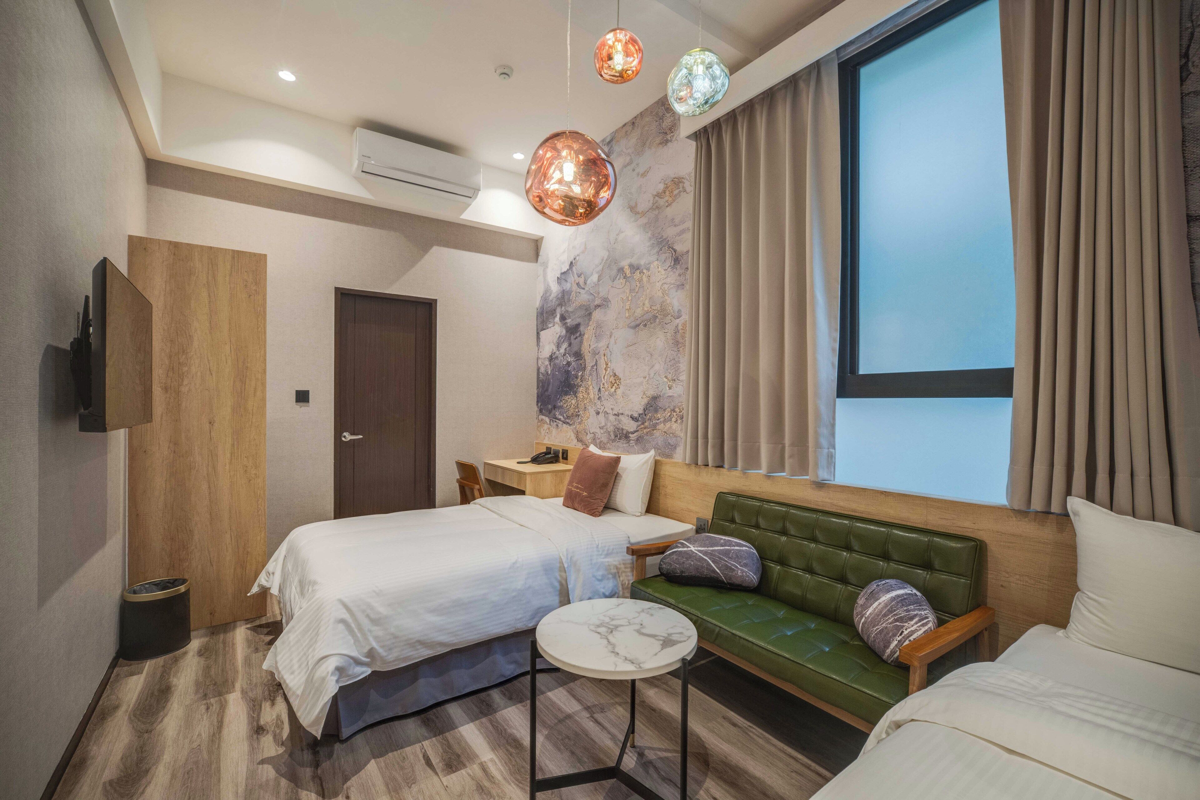 A modern hotel room with a large bed, a green sofa, a small round table, and a large window with curtains.
