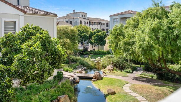 A serene courtyard with a small pond, lush greenery, and a pathway leading to a building with a tiled roof. - Bougain Villa CB206 (Cape Town)