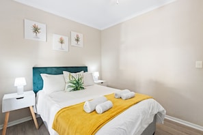 A neatly made bed with a yellow throw blanket and rolled towels, a teal headboard, white nightstands with lamps, and framed botanical prints on the wall.