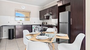 A modern kitchen with dark wooden cabinets, a stainless steel refrigerator, and a dining table set for four. - Bougain Villa CB206 (Cape Town)