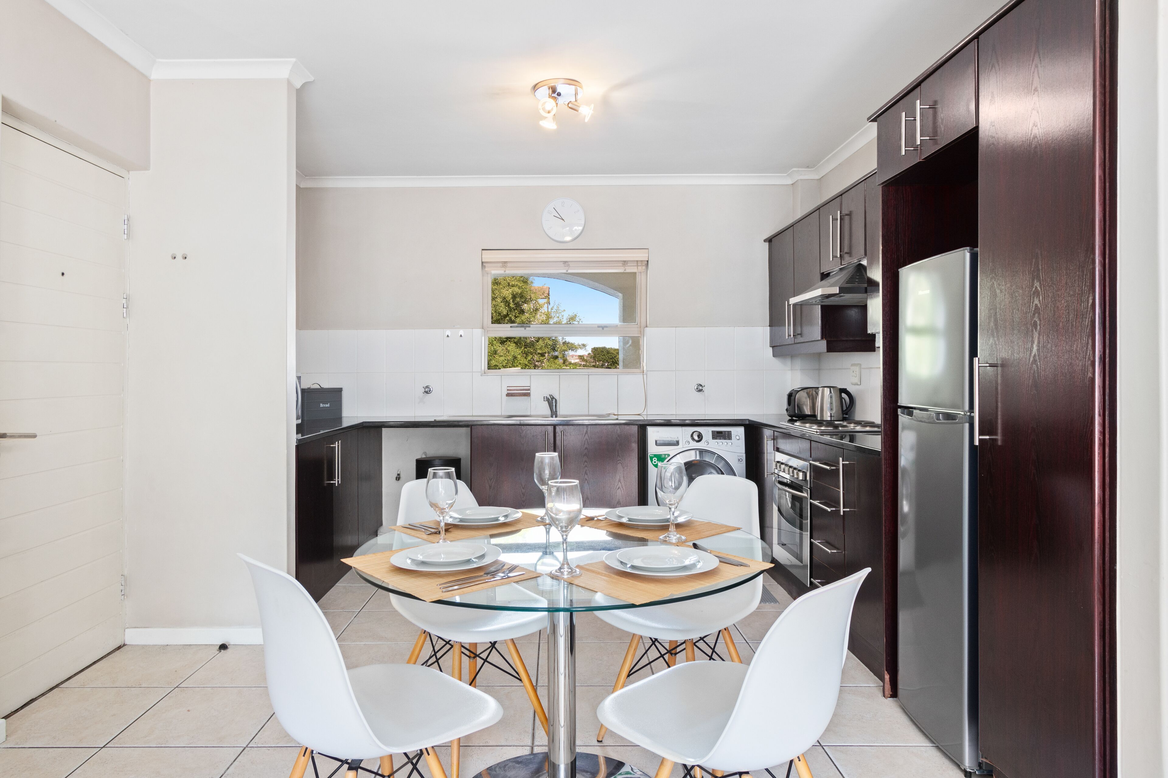 A modern kitchen with a glass dining table, white chairs, and dark wooden cabinets.