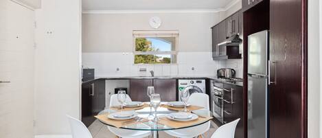 A modern kitchen with a glass dining table, white chairs, and dark wooden cabinets.