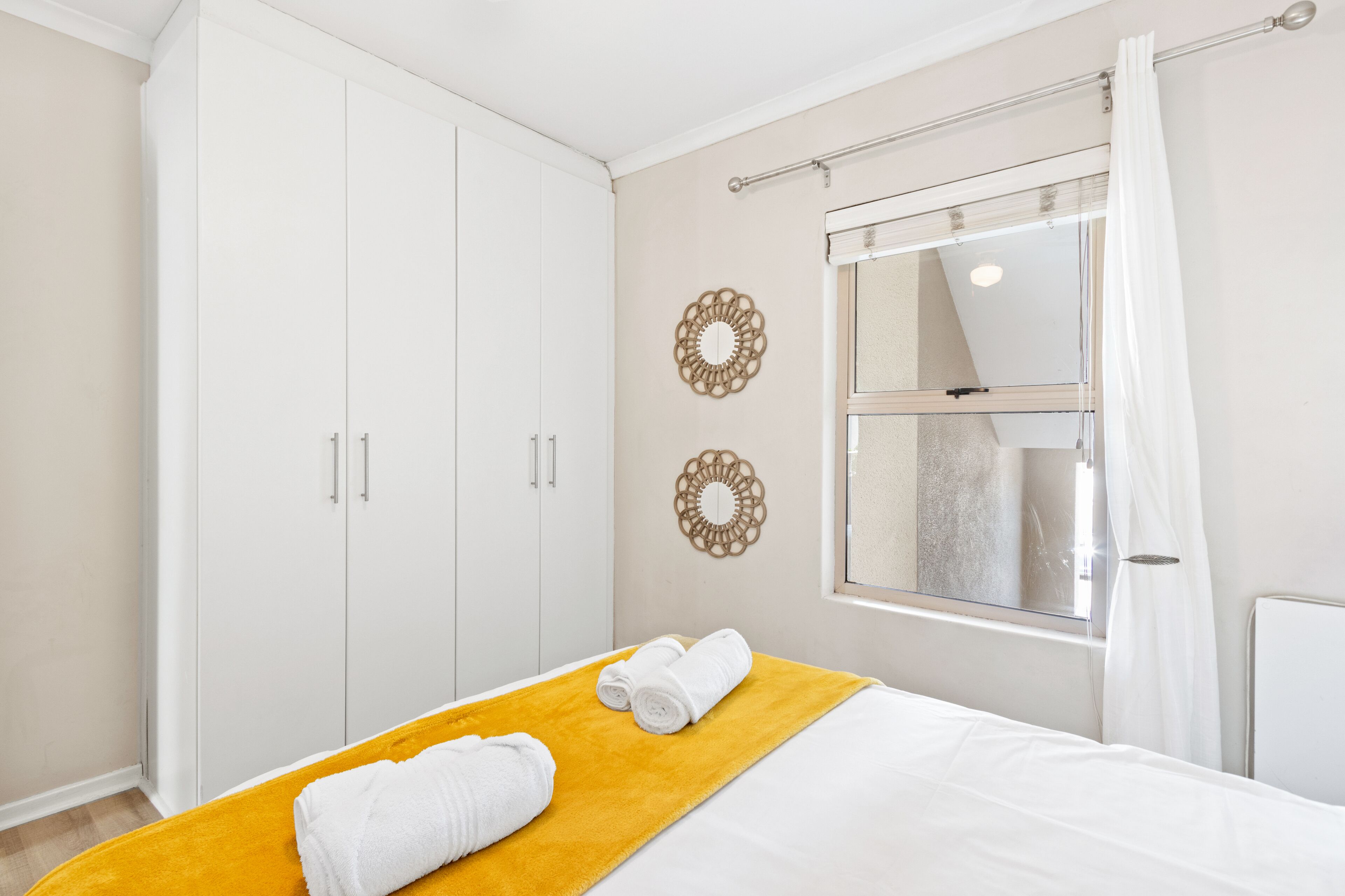 A bedroom with a bed, a yellow throw blanket, two white towels, a wardrobe, and decorative wall mirrors.