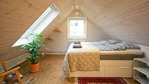 Une petite chambre mansardée, chaleureuse et accueillante, avec un plafond en pente, un puits de lumière, un lit recouvert d’une couette matelassée, une plante en pot et un radiateur.